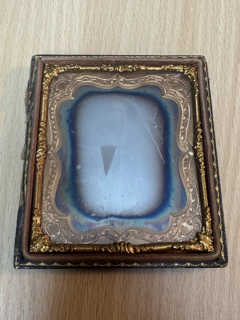Daguerréotype dans son étui de cuir et son cadre doré, vu de face sous un éclairage ordinaire : la plaque ressemble à un miroir terni, bleuté sur les bords, sans image lisible.