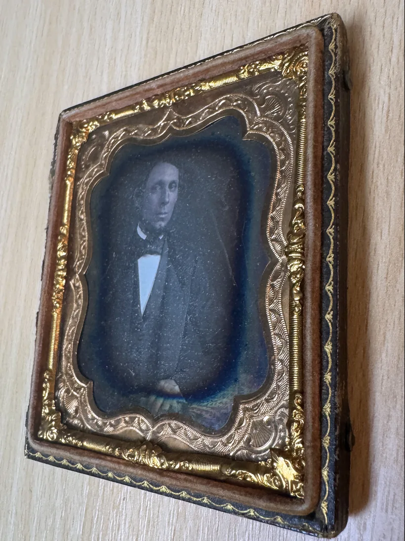 Daguerréotype dans son étui de cuir et son cadre doré, incliné : le portrait d'un homme en costume sombre et nœud papillon apparaît sur la plaque.
