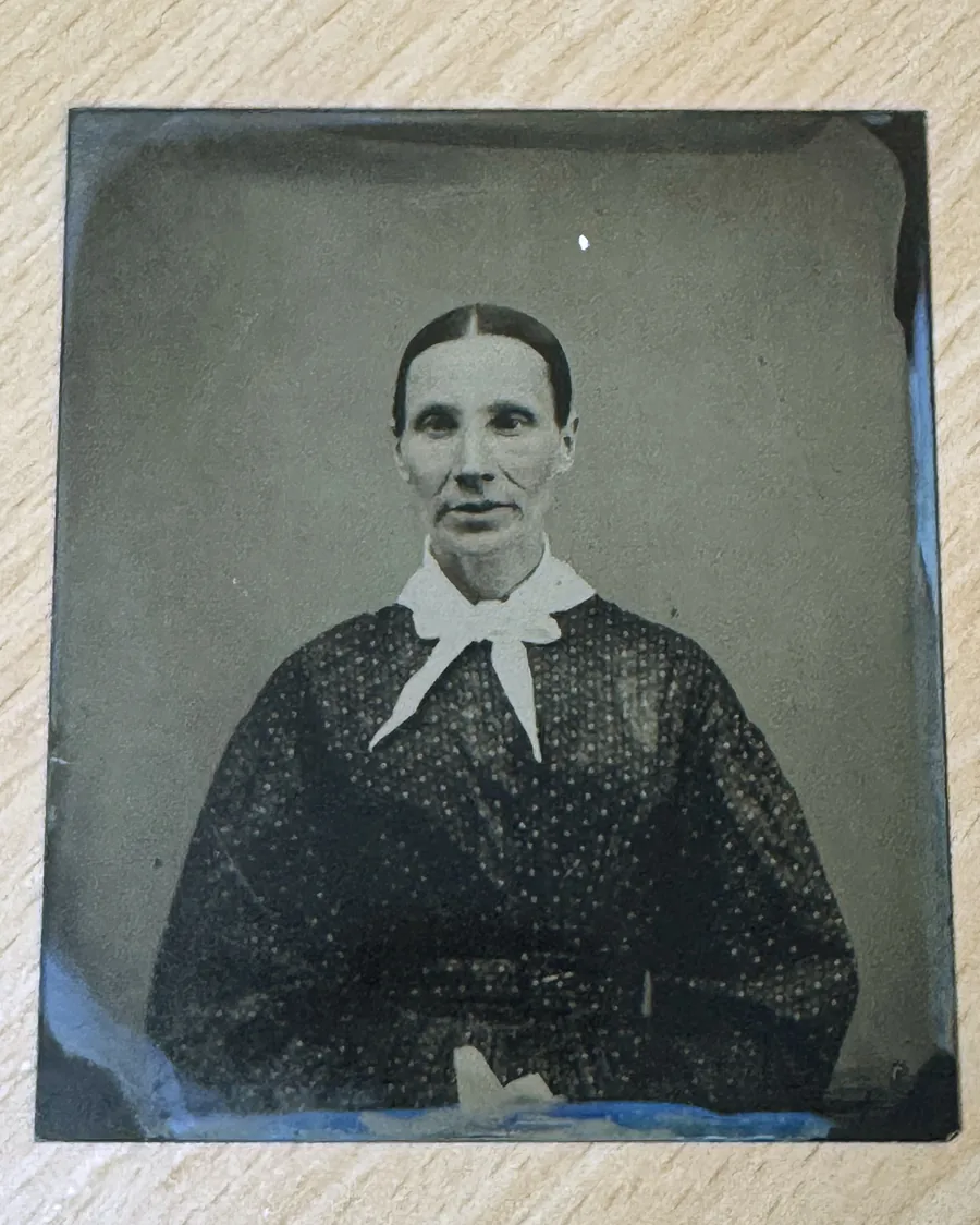 Ferrotype : portrait d'une femme aux cheveux séparés par une raie, col blanc noué, robe sombre à petits motifs, sur une plaque de fer laquée au fond gris.
